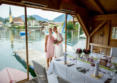 Braut und Gast posieren bei Hochzeit im Seehotel Malerwinkel, dekorierter Tisch, Aussicht auf Tegernsee im Hintergrund.