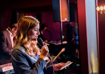 Frau haelt Rede bei Hochzeitsfeier mit Mikrofon. Hintergrund zeigt Musiker. Hochzeitsreportage in Bernried.