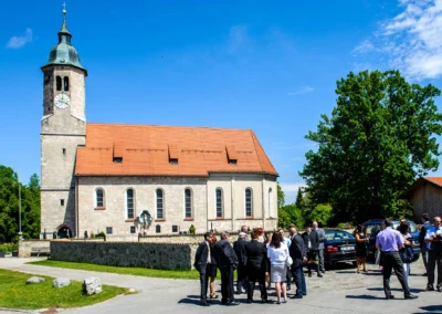 Gäste bei kirchlicher Trauung vor der Frauenkirche Osterwarngau. Hochzeitsfotografie in Oberbayern bei Sonnenschein.