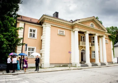 Personen in schicker Kleidung mit bunten Regenschirmen stehen vor klassischem Gebäude mit Säulen, grauer Himmel.
