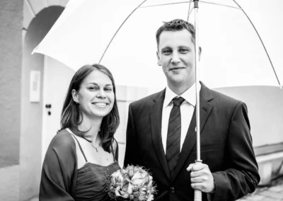 Paar in eleganter Kleidung unter weißem Regenschirm; Frau hält Blumenstrauß, lächelt; Mann trägt Anzug, steht ernst.