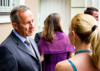 Ein Mann im Anzug spricht mit einer Frau im blauen Kleid; im Hintergrund weitere Personen, diffuses Licht.