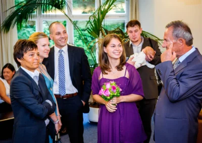 Mehrere Personen in formeller Kleidung stehen lächelnd zusammen; eine Frau hält einen Blumenstrauß, im Hintergrund ein Mann mit Baby.