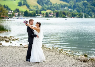 Brautpaar am Schliersee in Hochzeitskleidung am Ufer, umgeben von malerischer Landschaft. Hochzeitsfoto in Oberbayern.