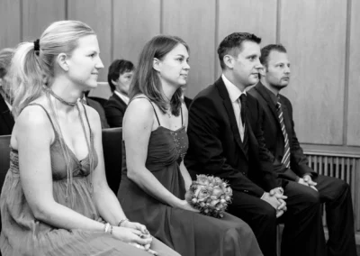 Vier Personen in formeller Kleidung sitzen nebeneinander auf einer Bank, zwei Frauen in Kleidern, zwei Männer in Anzügen.