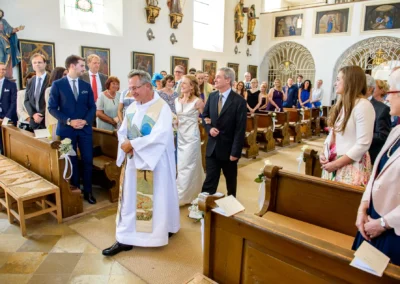 Priester fuehrt Brautpaar durch die Kirche St. Benedikt Gelting, Gaeste stehen klatschend bei kirchlicher Hochzeit.