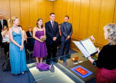 Personen in festlicher Kleidung stehen vor einer Standesbeamtin; eine Frau trägt ein lila Kleid und hält einen Blumenstrauß.