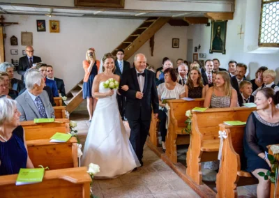 Braut schreitet mit Brautvater zur kirchlichen Trauung in Glonn. Hochzeitsreportage in einer Kirche mit Gästen.