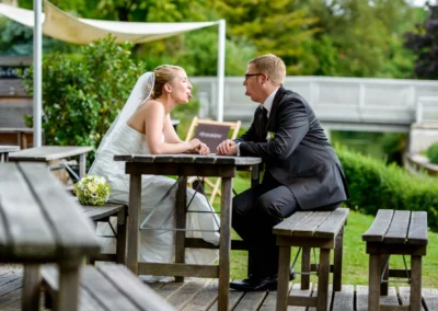 Brautpaar beim Shooting in einem Garten in Fürstenfeldbruck, lachend an Holztischen sitzend. Hochzeitsfotografie.