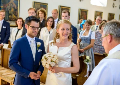 Brautpaar vor dem Priester während der kirchlichen Trauung in St. Benedikt Gelting, umgeben von Gästen.