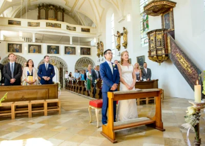 Brautpaar bei kirchlicher Trauung in St. Benedikt Gelting. Hochzeitsfoto zeigt Zeremonie mit Gaesten und Priester.