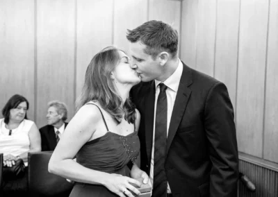 Ein Paar in festlicher Kleidung küsst sich innig in einem Raum mit Holzvertäfelung, im Hintergrund sitzen zwei weitere Personen.