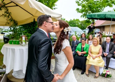 Brautpaarkuss bei freier Trauung, umgeben von Gaesten auf Schloss Sulzemoos. Hochzeitsfoto im Freien.