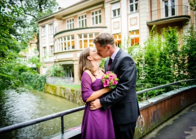 Paar in festlicher Kleidung küsst sich auf einer Brücke, Frau hält Blumenstrauß, im Hintergrund Gebäude und Fluss.