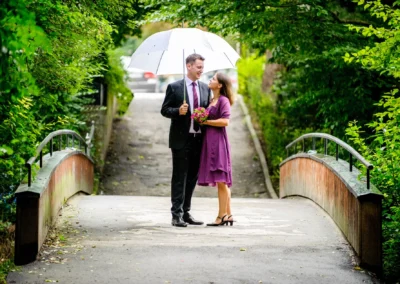 Ein Paar steht unter einem weißen Schirm auf einer Brücke, umgeben von üppigem Grün; sie hält einen Blumenstrauß.