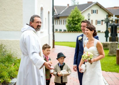 Brautpaar lacht mit Priester nach kirchlicher Trauung in Dorfkirche Moosach, Hochzeitsfoto in Oberbayern.