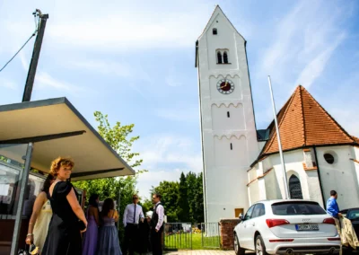 Gäste bei einer kirchlichen Trauung in Ottendichl, Oberbayern, neben einer Kirche und Autos geparkt.