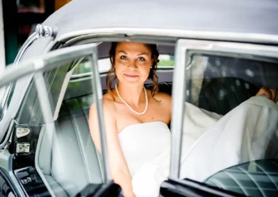 Eine lächelnde Braut in einem Auto, trägt ein schulterfreies Kleid und eine Perlenkette, sitzt auf einem grauen Ledersitz.