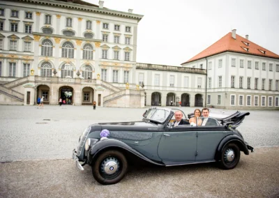 Ein altes Cabrio parkt vor einem großen, historischen Gebäude; drei Personen sitzen lächelnd im Auto.