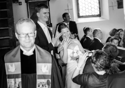 Brautpaar bei kirchlicher Trauung in Gut Georgenberg, lachend, mit anwesendem Pfarrer und Gaesten im Hintergrund.