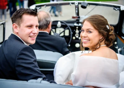 Lächelndes Brautpaar im offenen Oldtimer; die Frau im weißen Kleid, mit Perlenkette, der Mann im dunklen Anzug.