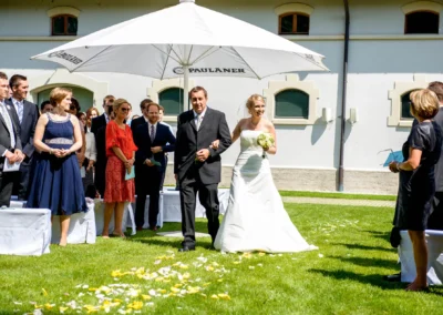 Braut schreitet mit Vater zur freien Trauung im Garten in Fuerstenfeldbruck, Gaeste im Hintergrund, Hochzeitsfoto.