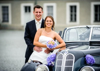 Brautpaar steht glücklich vor einem Oldtimer, die Braut hält einen Blumenstrauß, das Auto ist mit lila Blumen geschmückt.