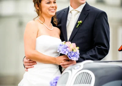 Brautpaar lächelt neben einem Oldtimer; die Braut trägt ein weißes Kleid mit Perlenkette, der Bräutigam einen dunklen Anzug.