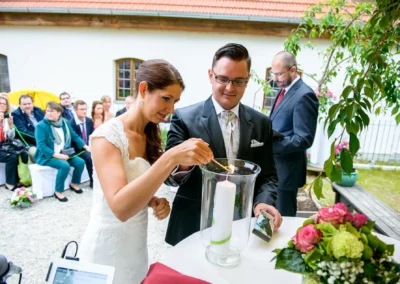Brautpaar entzuendet Kerze bei der freien Trauung auf Gut Schloss Sulzemoos, umgeben von Gaesten und Blumenarrangement.