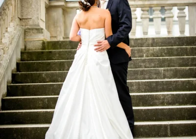 Brautpaar auf einer Treppe, die Braut im weißen Kleid umarmt den Bräutigam im dunklen Anzug; barocker Balkon im Hintergrund.