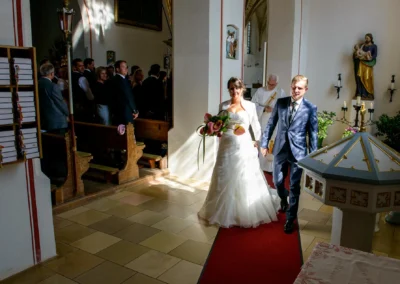 Brautpaar beim Kirchenauszug in St. Vitus, begleitet von einem Priester. Hochzeitsfotograf in der Kirche in Oberbayern.