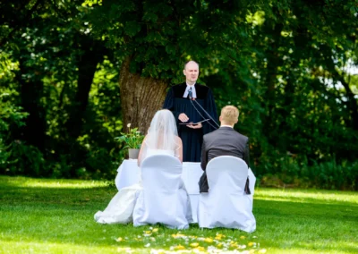 Freie Trauung im Freien, Hochzeitsfotografie in Kloster Fuerstenfeld, Paar und Redner vor gruenem Hintergrund.