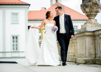 Brautpaar spaziert lächelnd auf einer Terrasse; die Braut trägt ein weißes Kleid und einen Blumenstrauß, der Bräutigam einen schwarzen Anzug.