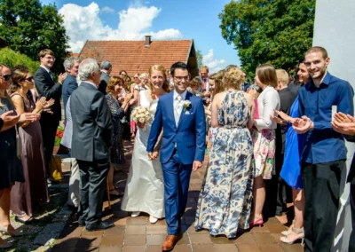 Brautpaar verlässt Kirche bei Hochzeit in St. Benedikt Gelting, umgeben von applaudierenden Gästen im Freien.