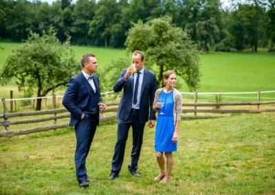 Drei Hochzeitsgaeste stehen im Gruenen bei Gut Georgenberg und trinken, im Hintergrund eine Holzumzäunung.