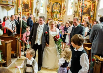 Brautpaar bei kirchlicher Trauung in Warngau, umgeben von Gästen in traditioneller Kleidung. Hochzeitsfoto Bayern.