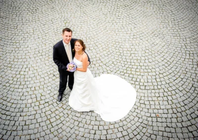 Brautpaar lächelt nach oben auf gepflastertem Platz; Bräutigam trägt Anzug, Braut hält Brautstrauß, langes weißes Kleid.