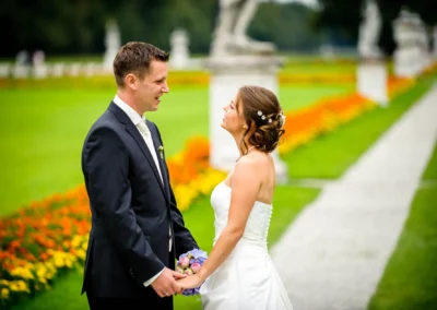 Brautpaar hält Hände in einem farbenfrohen Garten, sie trägt ein weißes Kleid, er einen dunklen Anzug, beide lächeln sich an.
