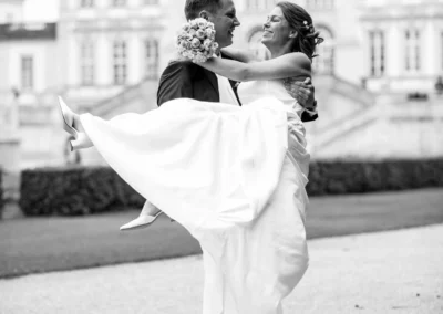 Das Brautpaar lacht; er hebt sie hoch, sie hält einen Brautstrauß. Im Hintergrund ein großes, elegantes Gebäude.