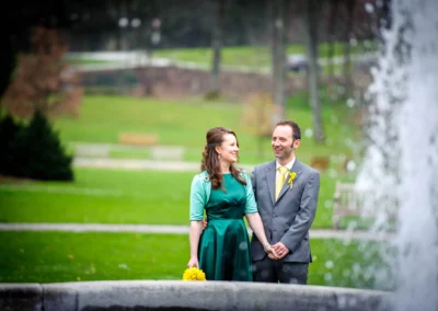 Brautpaar im Kurpark Bad Tölz laechelnd beim Hochzeitsportrait neben einem Brunnen, festgehalten in der Hochzeitsfotografie.