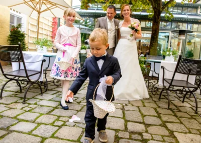 Kind streut Blumen bei Hochzeit im Gasthof Stangl. Brautpaar im Hintergrund laechelt. Hochzeitsfoto fuer Reportage.