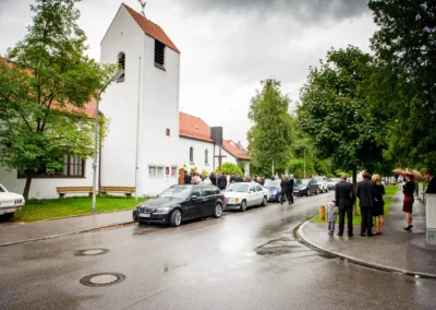 Menschen in feiner Kleidung stehen vor einer weißen Kirche mit rotem Dach; Allee von Bäumen, nasse Straße, parkende Autos.