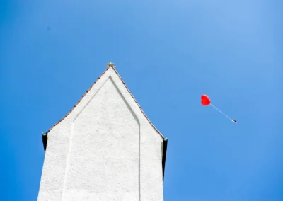 Kirchturm mit rotem Herzballon am Himmel, Hochzeit in Ottendichl bei München, festgehalten in einer Hochzeitsreportage.