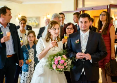 Brautpaar mit Blumenstrauss bei Hochzeitsfeier in Grosshartpenning, umgeben von Gaesten mit Getraenken. Hochzeitsfoto.