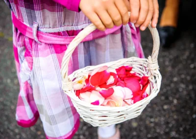 Kind mit kariertem Kleid hält weißen Korb voller bunter Rosenblätter auf dunklem Boden.