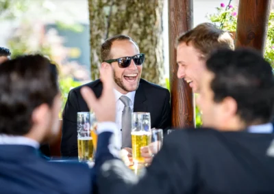 Männer in Anzügen lachen bei einer Hochzeit im Gasthof Widmann in Oberbayern, im Freien bei Getränken. Hochzeitsreportage.