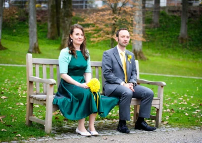 Brautpaar sitzt auf einer Holzbank im Kurpark Bad Tölz, Hochzeitsportraits im Freien, festliche Kleidung.