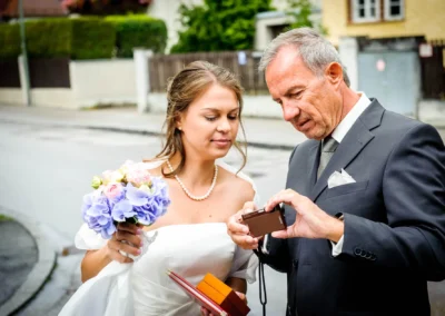 Braut mit Blumenstrauß und älterer Mann im Anzug betrachten eine Kamera auf einer Straße.