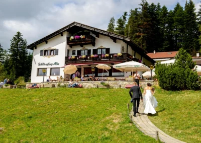 Brautpaar bei Hochzeit auf Schliersberg Alm, Oberbayern, geht auf Gasthaus zu, umgeben von gruenen Wiesen und Gaesten.
