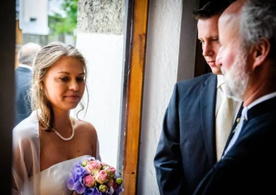 Braut mit Blumenstrauß und Perlenkette neben zwei Männern in Anzügen, in dämmriger Innenlichtstimmung, konzentrierter Blick.